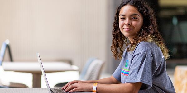 一名女子穿着印有Alamo Promise字样的灰色t恤，在笔记本电脑前工作
